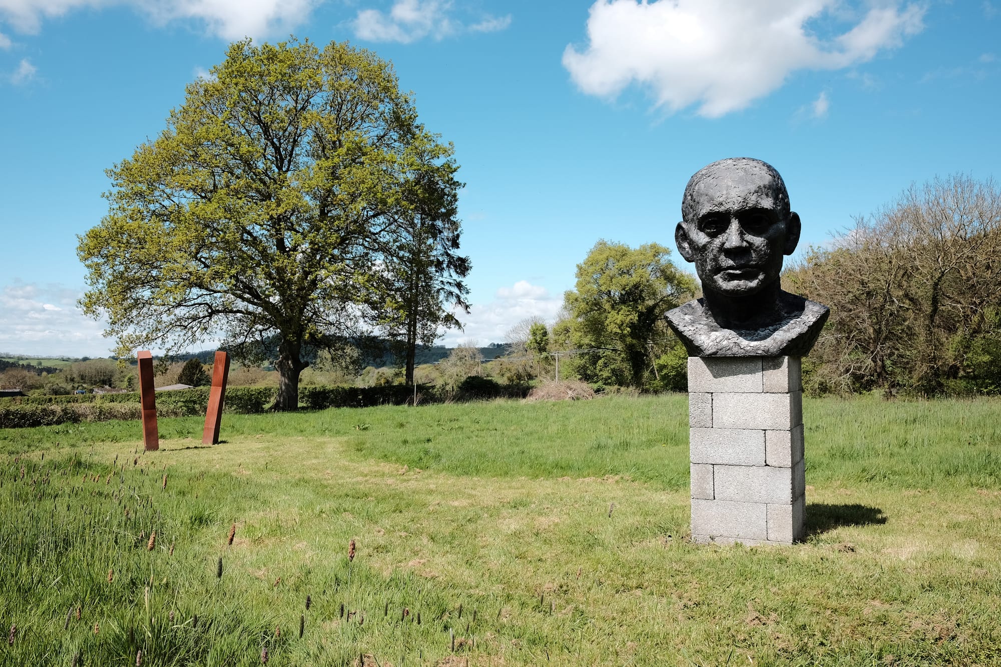 Two sculptures sit in a field of mown green grass: one comprised of brown rusty geometric shapes, the other a grey bust of a man's head