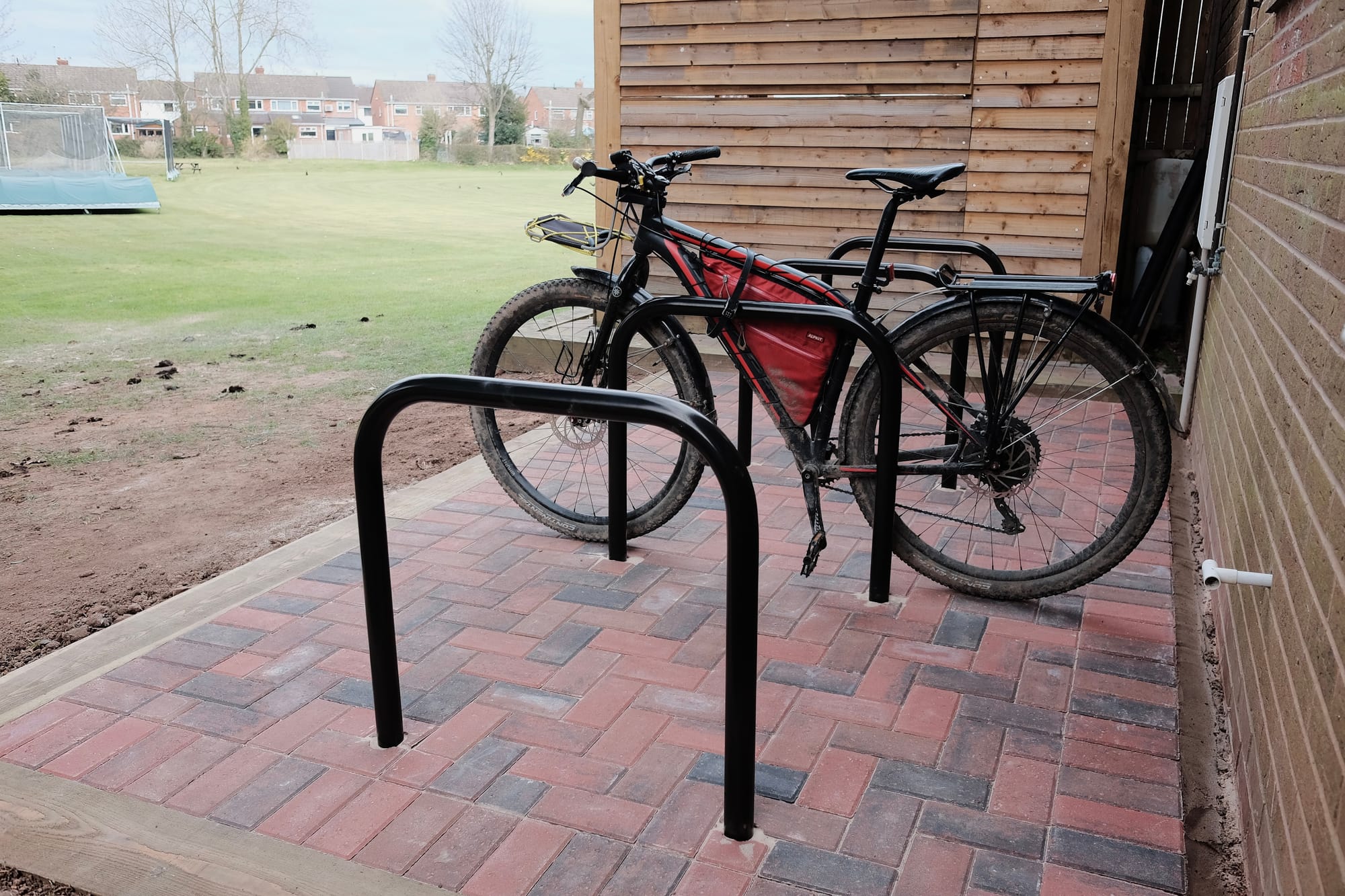 A black and red bike parked in a row of black stands in a red and blue brick base by the side of a grassy cricket field, with red brick houses behind. 