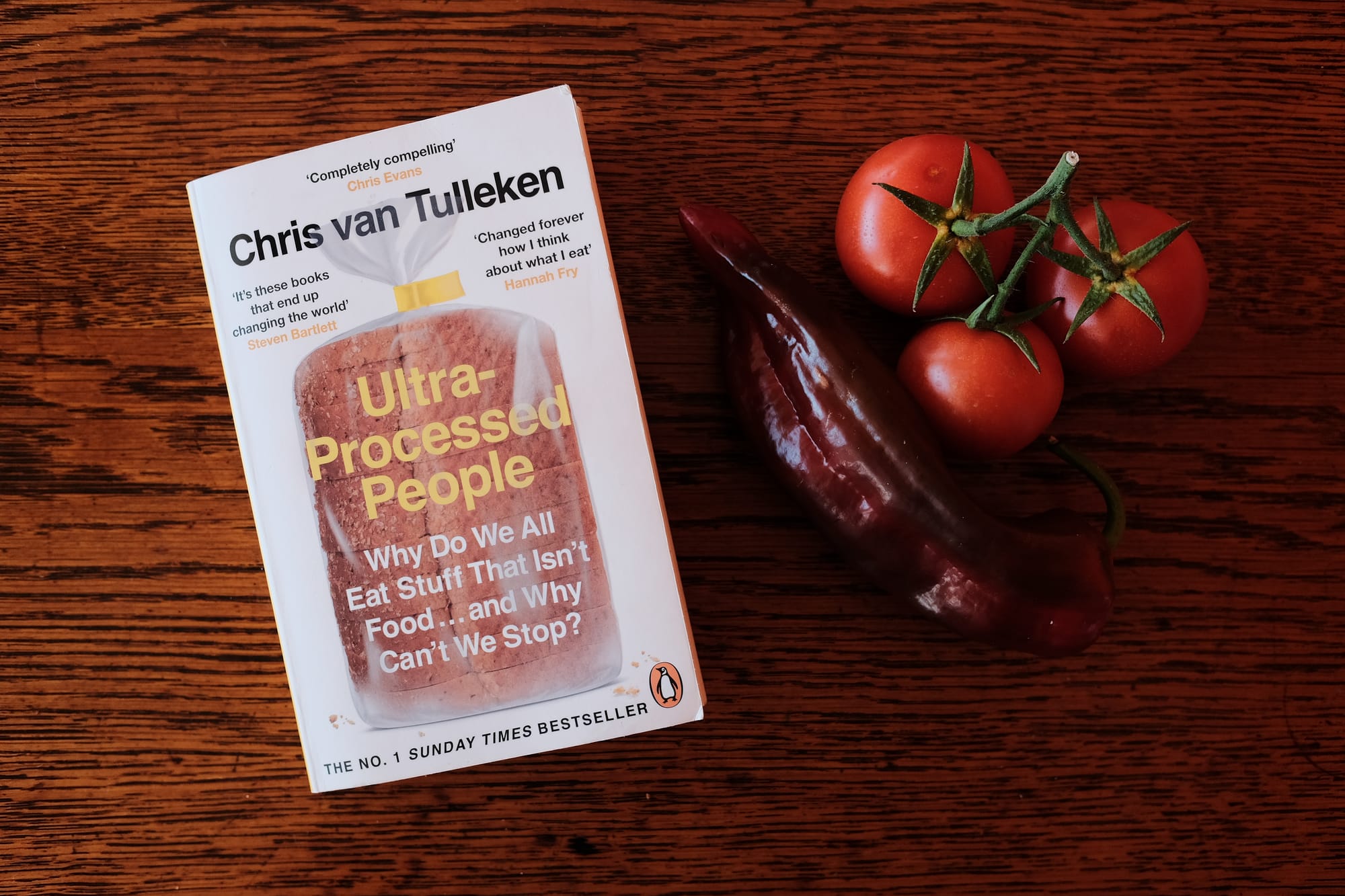 A copy of the book Ultra-Processed People by Chris van Tulleken sits on a wooden table next to a red pepper and red tomatoes.