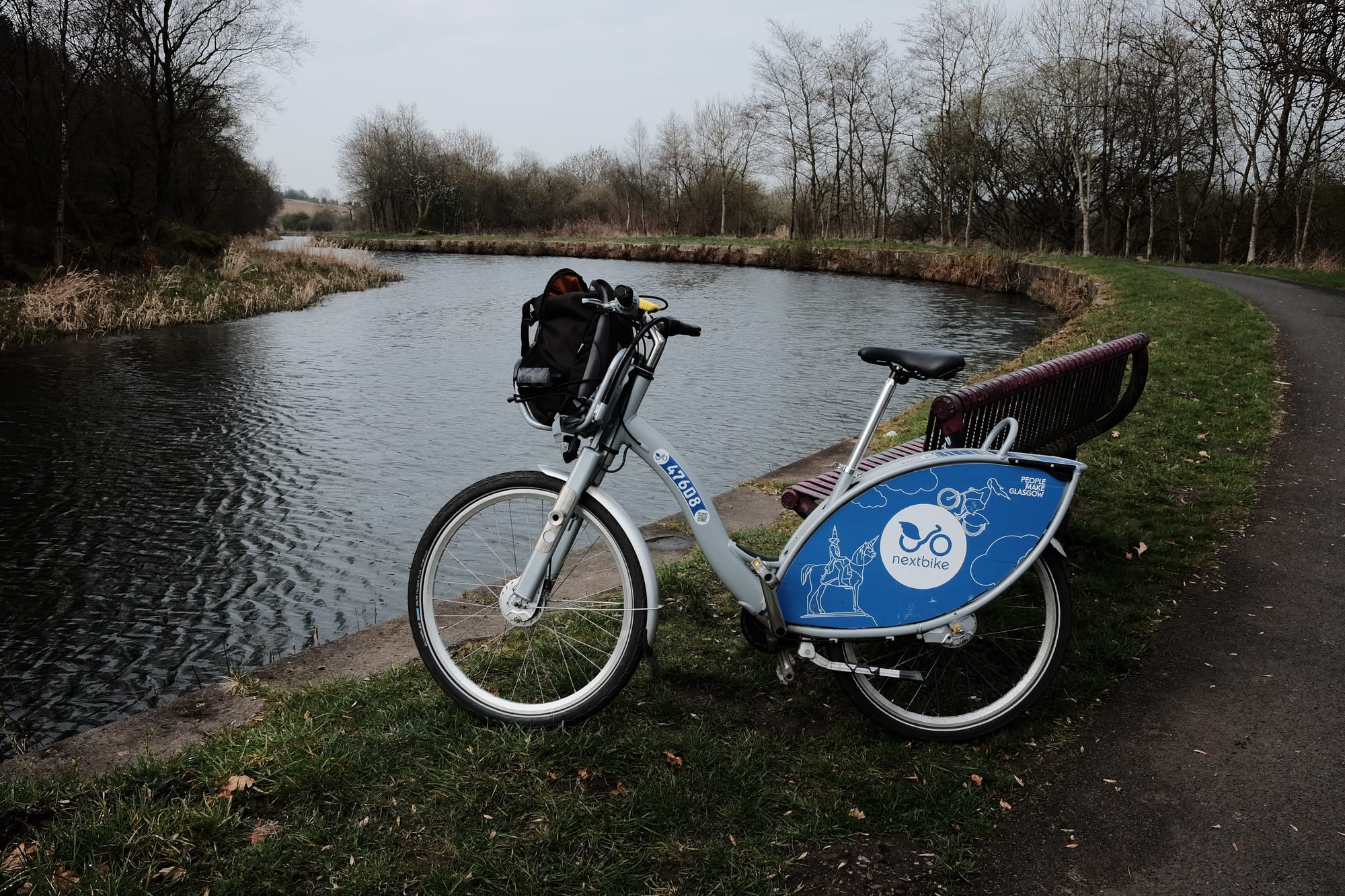 A blue and grey Nextbike leans against a red bench on the Forth and Clyde canal