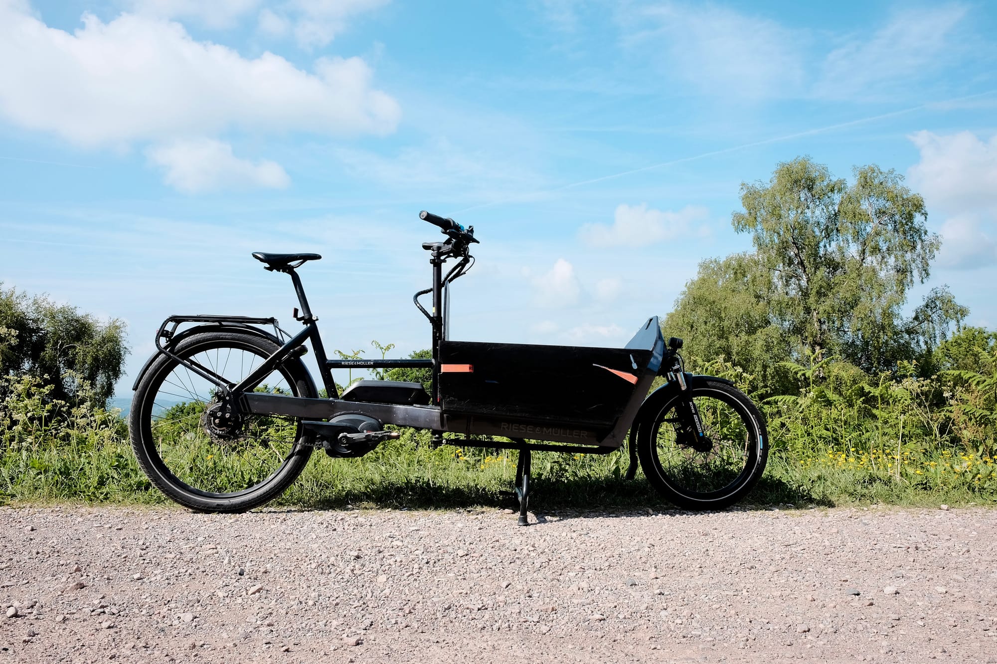 A black cargo bike stands on a gravel track with greenery and blue sky in the background.