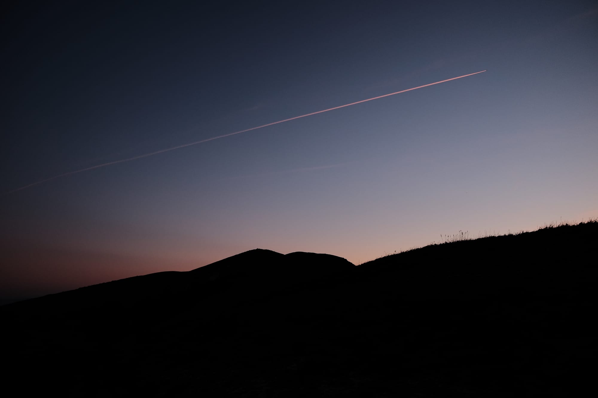 The silhouette of hills against a pink sunset, with the trail of a jet aeroplace above them.