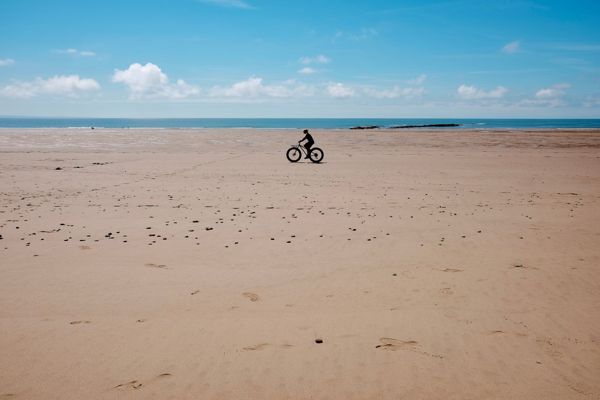 A lone cyclist on a fat bike rides on an empty sandy beach beneath a blue sky.
