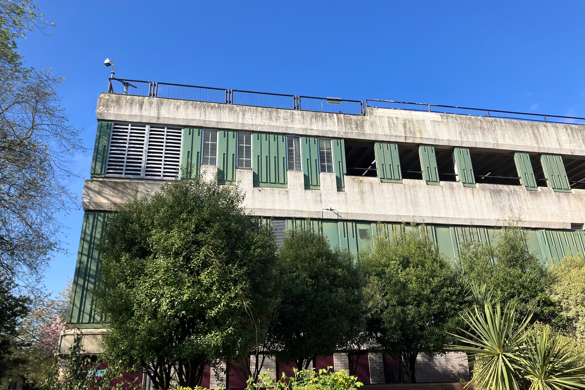 The grey and green structure of a multistorey car park is partially obscured by a line of small trees.