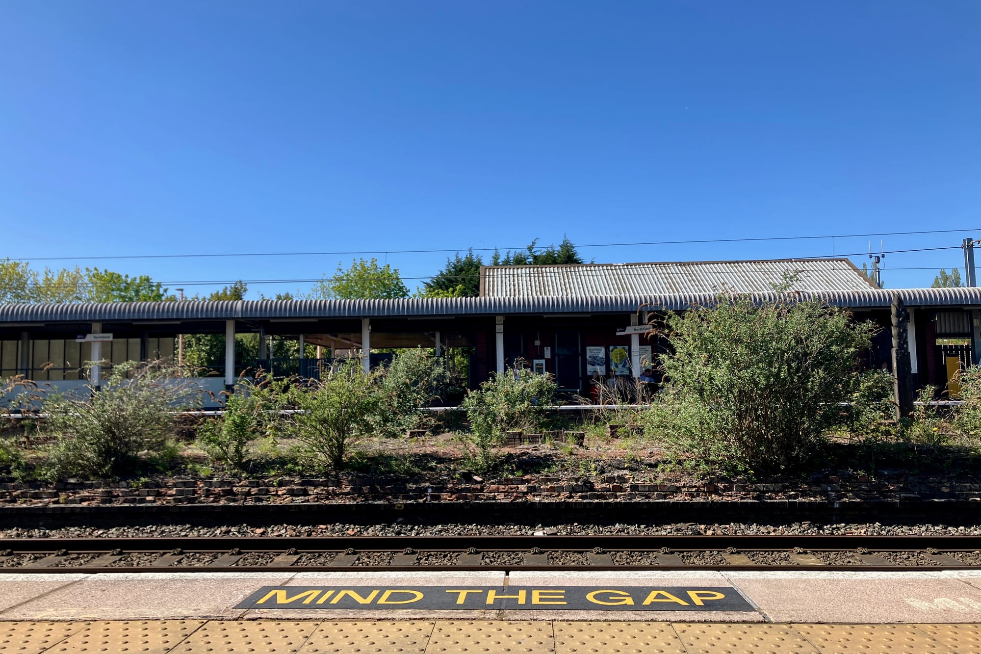 A view of Northfield Station in Birmingham with the central platform overgrown with shrubs and a 'mind the gap sign' visible in the foreground.