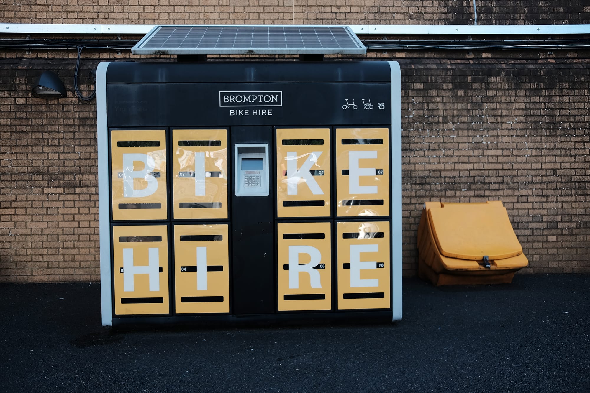 Yellow Brompton bike hire lockers stand against a yellow brick wall, with a yellow grit bin to the right of them.
