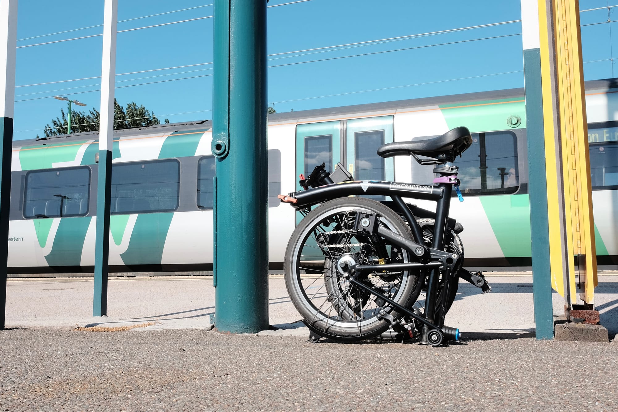 A Brompton bike sits folded on a station platform, a green and white train in the background and green poles either side.