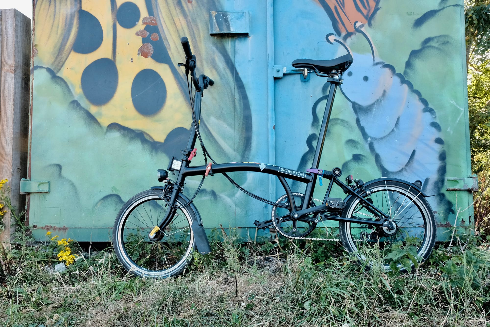 A black Brompton bike stands against a large cabinet with street art of creatures on it.