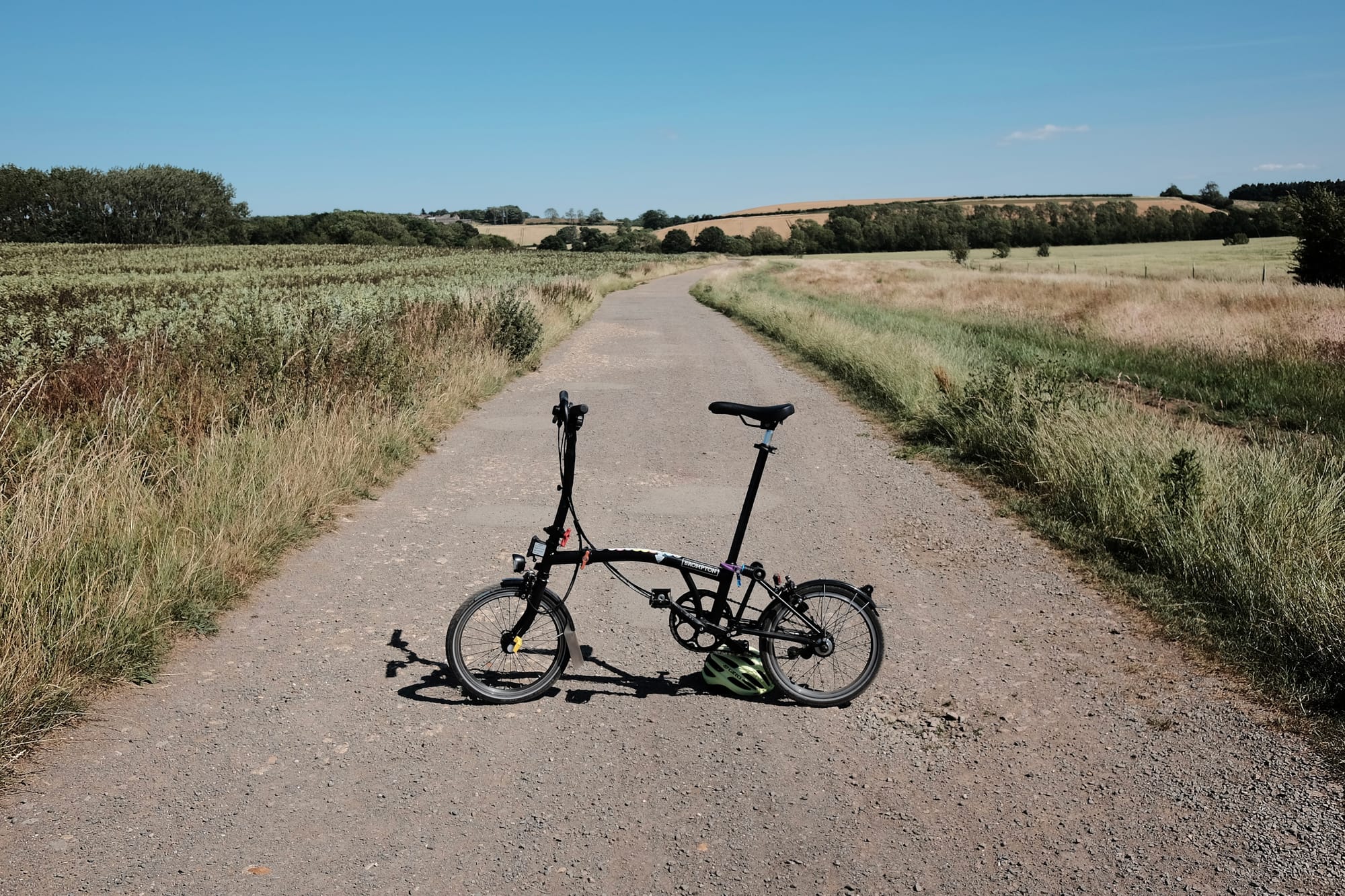 A Bromton bike stands in the middle of a stony byway propped up on a bike helmet, dry grassland all around.