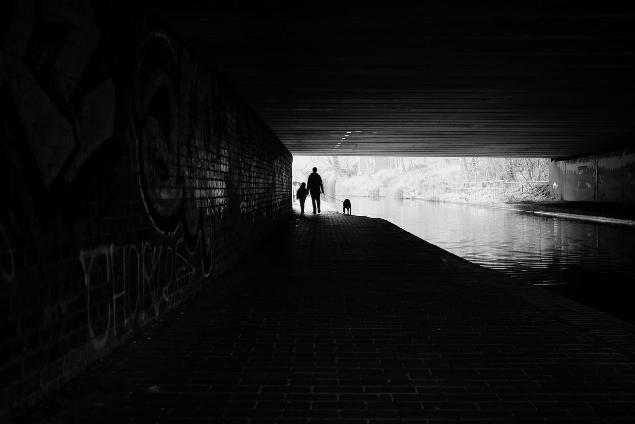 The silhouette of an adult, a child and a dog on a lead walking on the canal towpath underneath a road bridge.