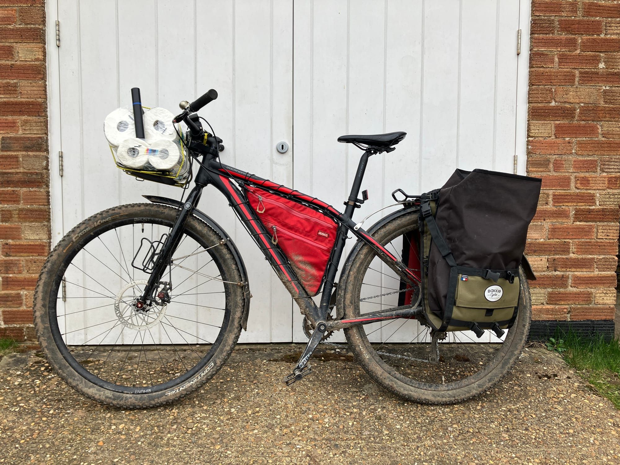 A black and red mounatin bike with panniers leans on white garage doors, its front rack loaded with a pack of toilet rolls