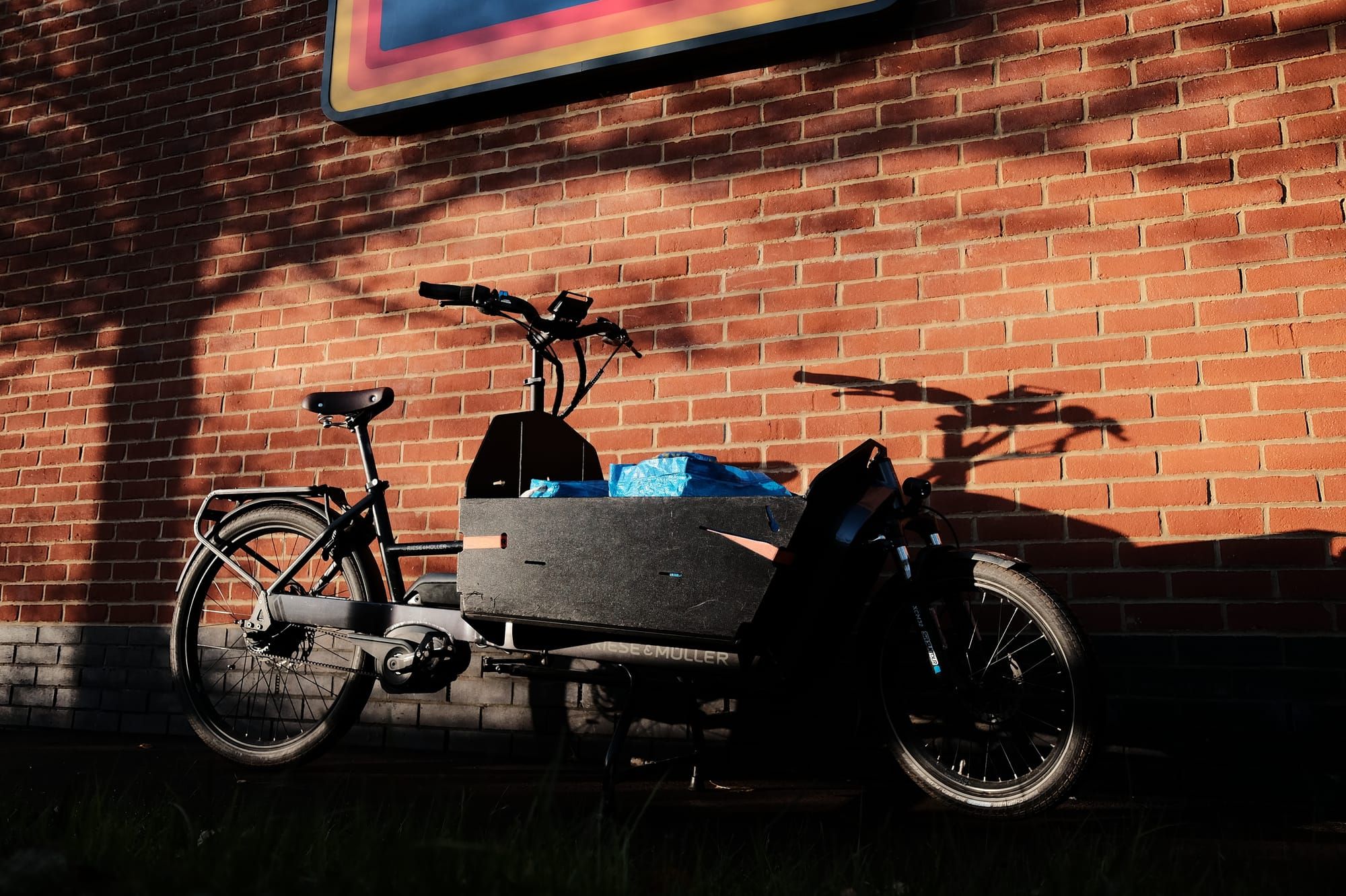 A black cargo bike leans against a brick wall, the strong sunlight casting shadows.