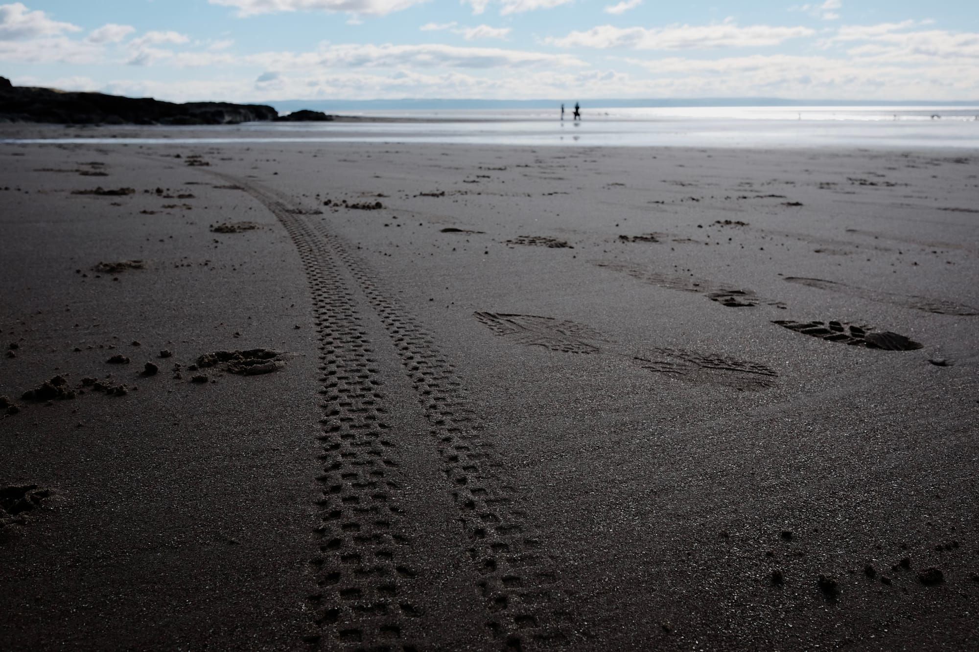 Bike tyre tracks and footprints are visible on a sandy beach, with the silhouettes of people in the distance by the sea.