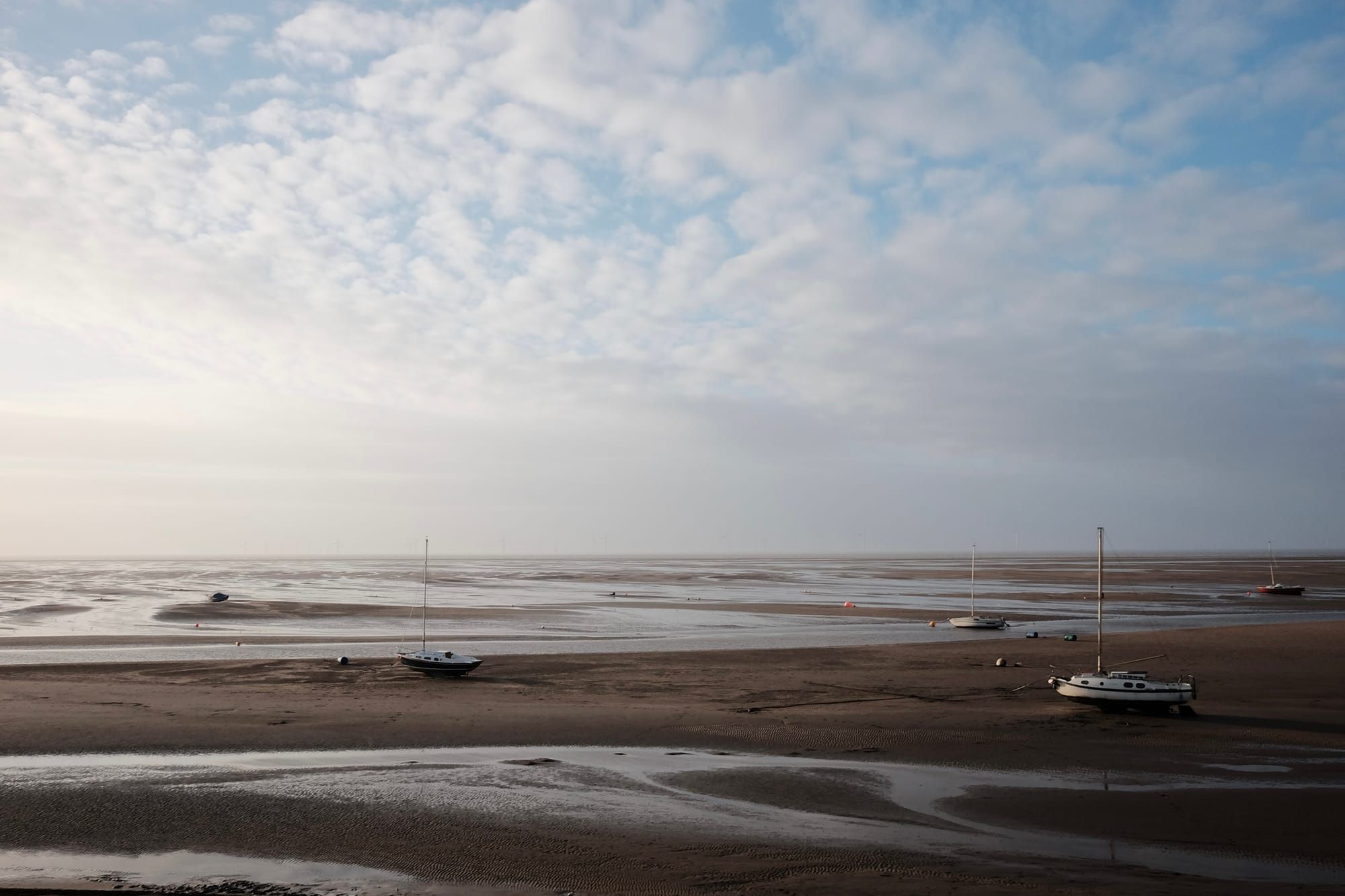A large expanse of sandy beach at low tide, with small sailing boats standing among channels of water.