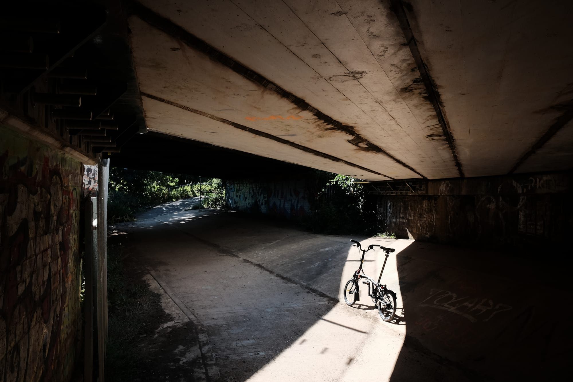 A Brompton bike stands in a shaft of sunlight in a concrete underpass.