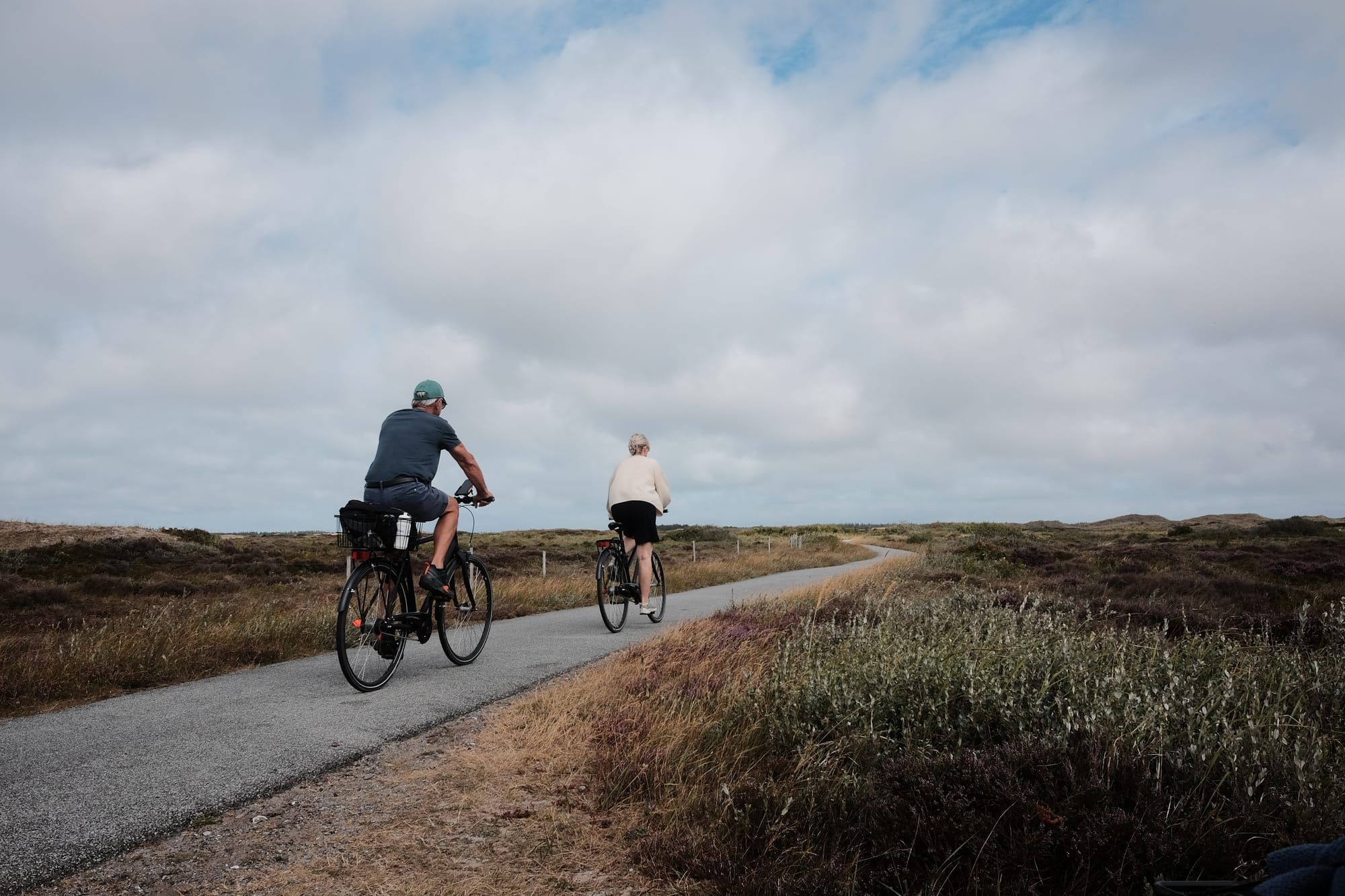 Two cyclists ride down a tarmac bike pathe in a heathland full of purple heather.