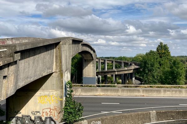 A grey and grubby concrete cycling and walking bridge spirals over a motorway and enters a green woodland.
