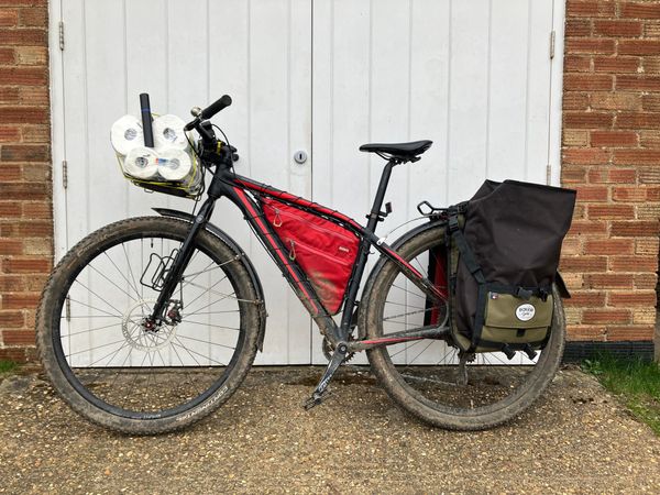 A black and red mounatin bike with panniers leans on white garage doors, its front rack loaded with a pack of toilet rolls