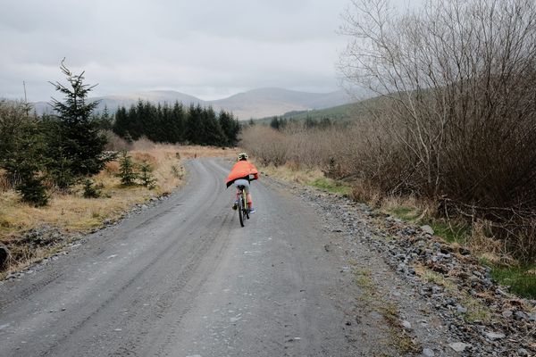 A child in a billowing organge jacket cycles away down a sweeping forest track, mountains in the distance.