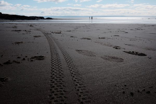 Bike tyre tracks and footprints are visible on a sandy beach, with the silhouettes of people in the distance by the sea.