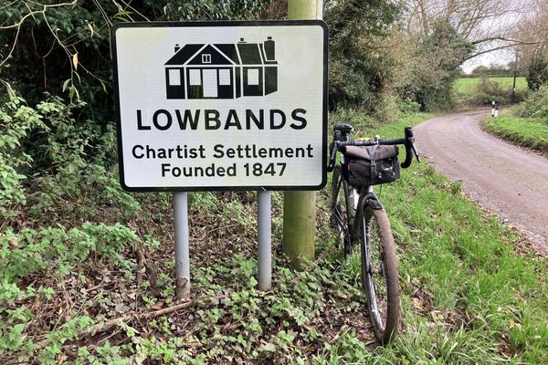 Along a tree-lined coutnry lane, a gravel bike with a black handlebar bag leans on a road sign with the words: "Lowbands: Chartist Settlement Founded 1847"