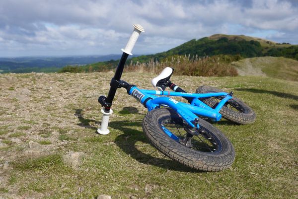 A blue Hoy balance bike rests on its side atop a grassy hill, with a cloudy sky in the background.