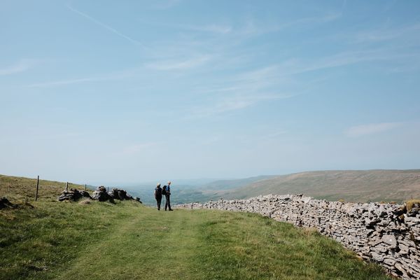 Two people walk along a high, grassy track bordered by a drystone wall. Beyond them is a low valley, above them only sky.