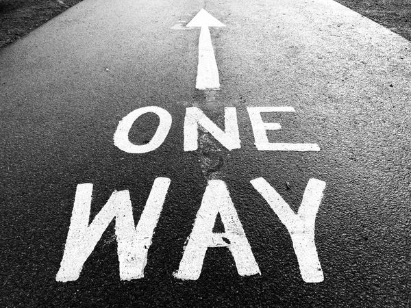 A black and white photo of a road with 'one way' painted on it, with an arrow pointing away from the camera position.