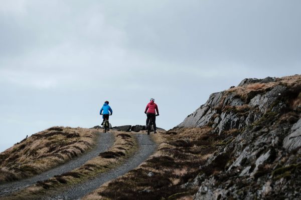 An adult cyclist in pink and child cyclist in blue pause at the summit of a mountain track.
