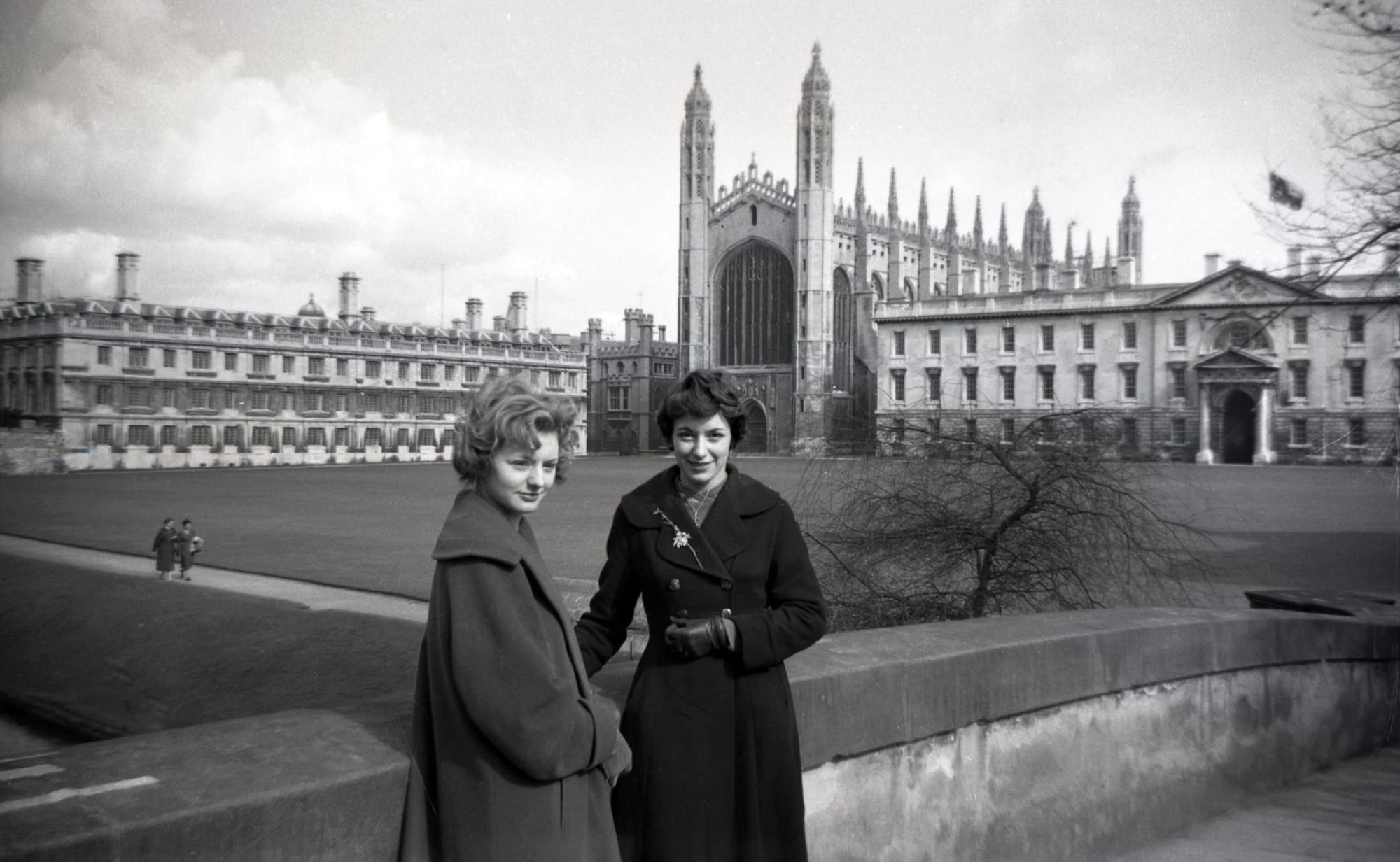  The image is a black-and-white photograph featuring two women standing in front of a historic building, likely a university or college. The architecture is grand and Gothic, with a large chapel-like structure visible in the background. The women are dressed in mid-20th century clothing, including long coats and gloves, suggesting the photo was taken during a cooler season. The scene has an academic or formal atmosphere, with the women standing on a stone bridge or walkway, and the expansive courtyard and buildings adding to the stately ambiance. The overall mood of the photograph is serene and timeless, capturing a moment of stillness in a historical setting.