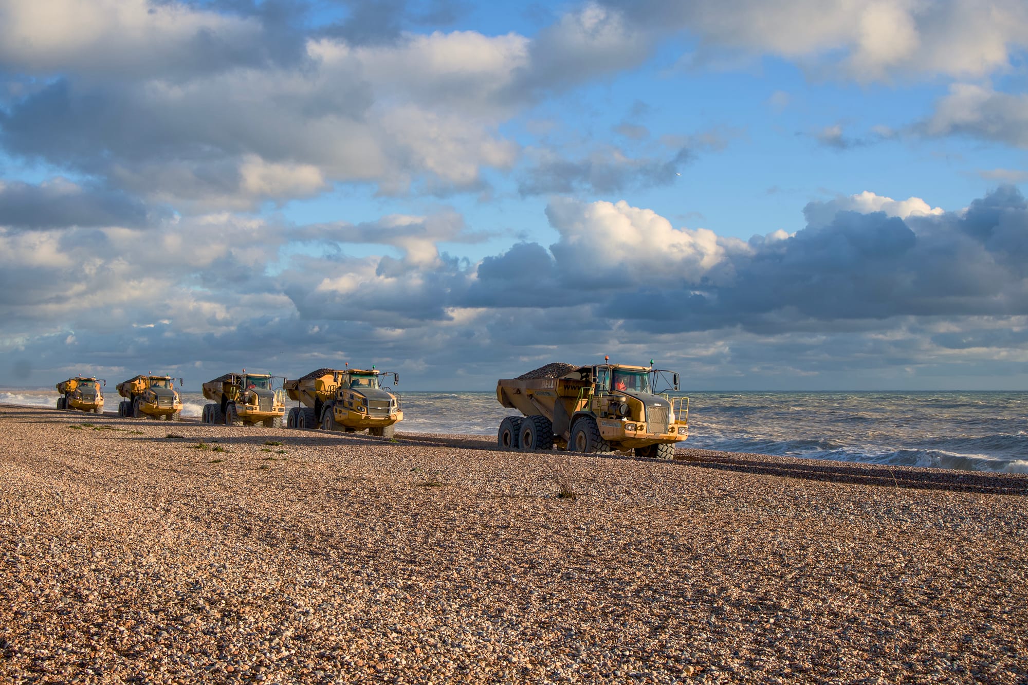 A row of large yellow dump trucks loaded with gravel lined up on a pebble beach, with the ocean and a dramatic cloudy sky in the background.