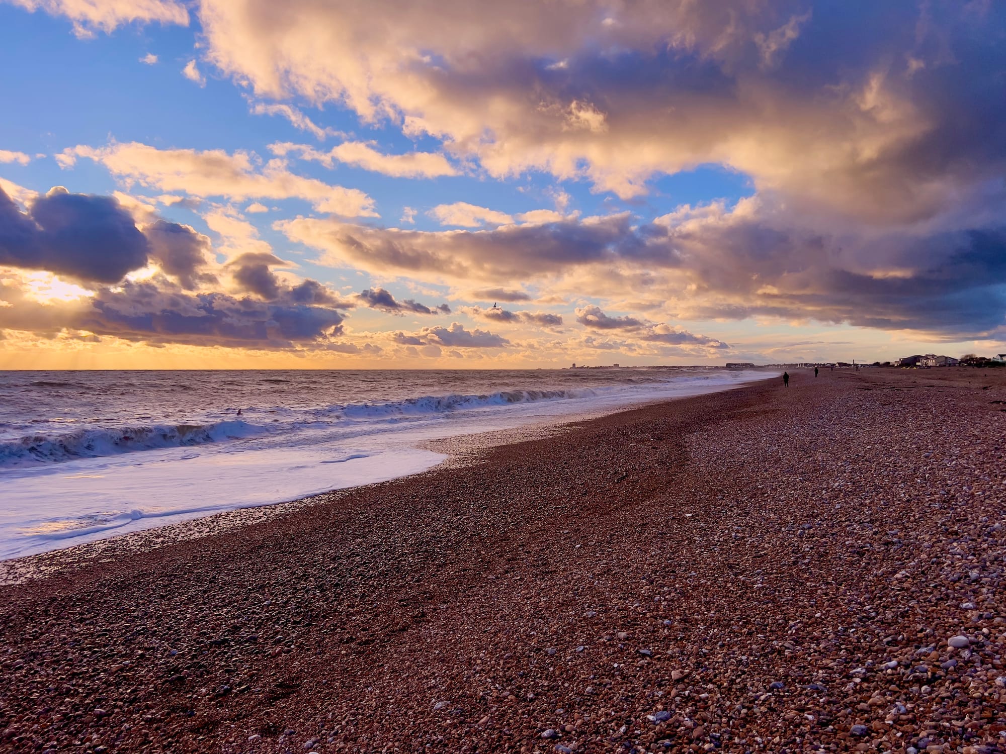 Pebble beach at sunset with waves rolling onto the shore, a vibrant sky filled with dramatic clouds and golden sunlight, and a few distant figures walking along the coastline.