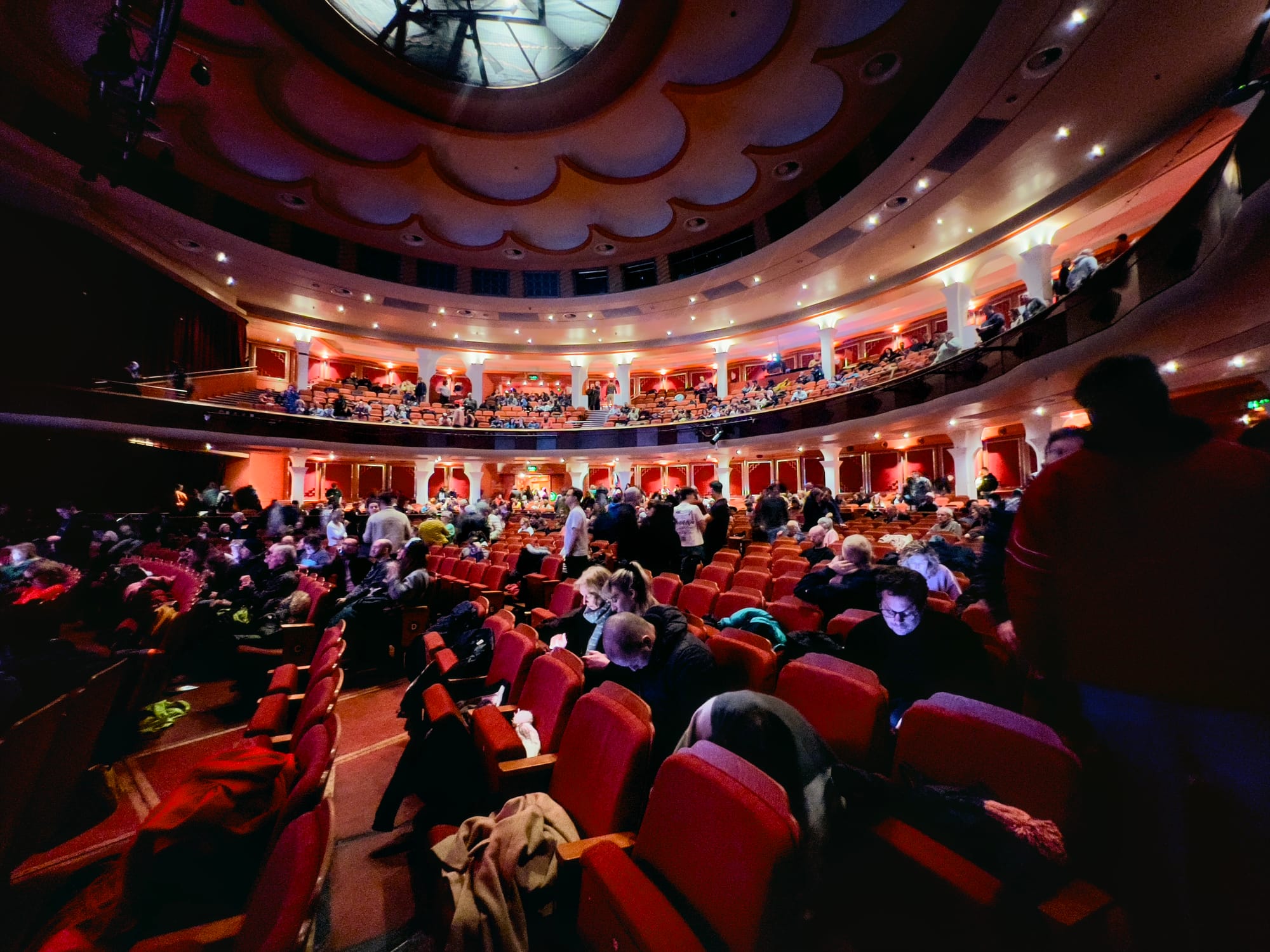 A theater auditorium filled with people before a performance. The image captures a grand, multi-tiered venue with red upholstered seats, ornate architectural details, and a large circular ceiling design. Audience members are seen chatting, finding their seats, and using their phones. Warm lighting illuminates the space, creating a lively and anticipatory atmosphere. The perspective is slightly tilted, adding a dynamic feel to the scene.