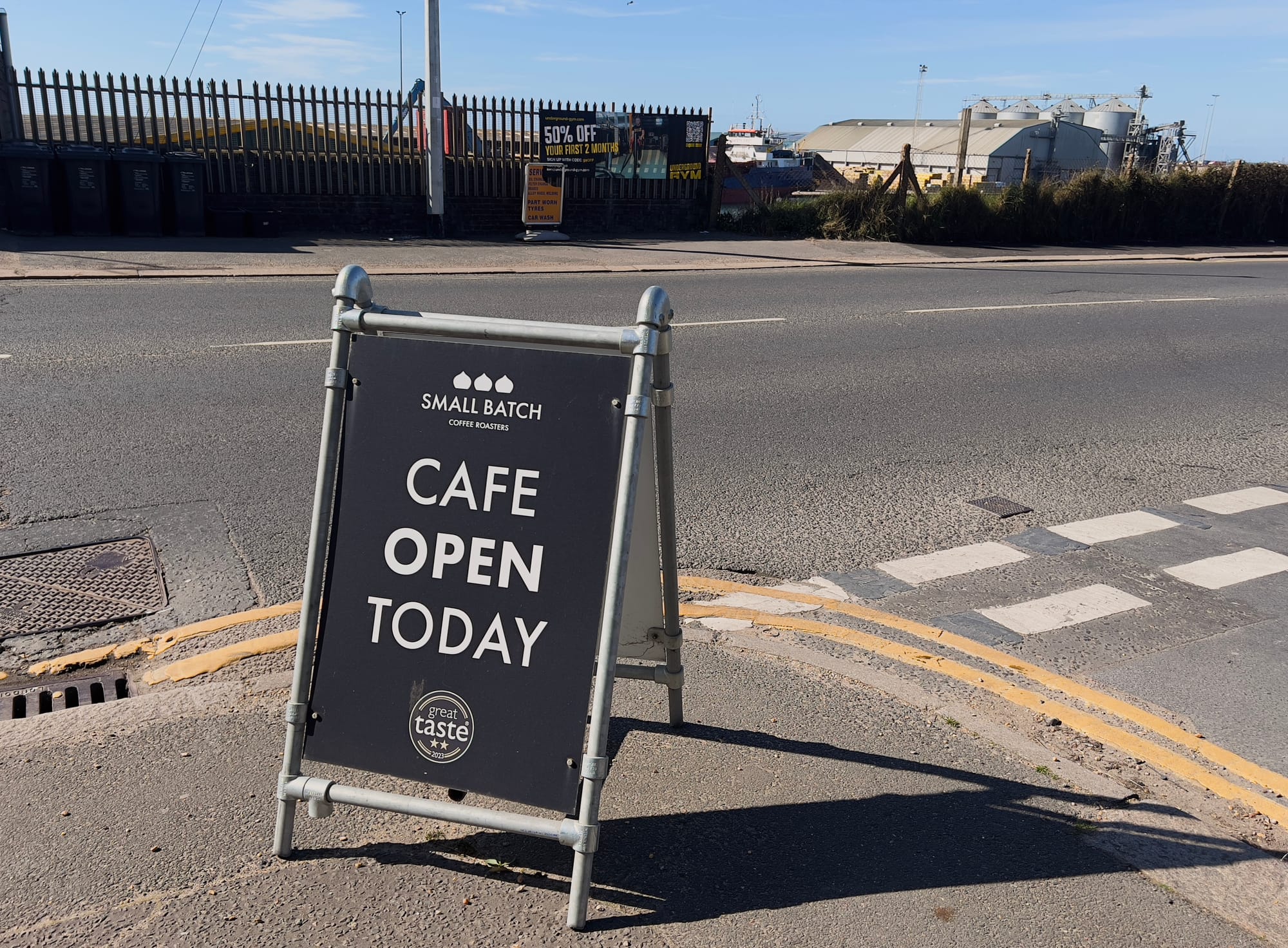 A sidewalk signboard for “Small Batch Coffee Roasters” stands near a crosswalk, displaying the message “Cafe Open Today” in bold white letters on a black background. The sign also features a “Great Taste” award logo. In the background, there is a road, a fence, an industrial area with large storage buildings, and a sign offering “50% off your first 2 months” at a gym.
