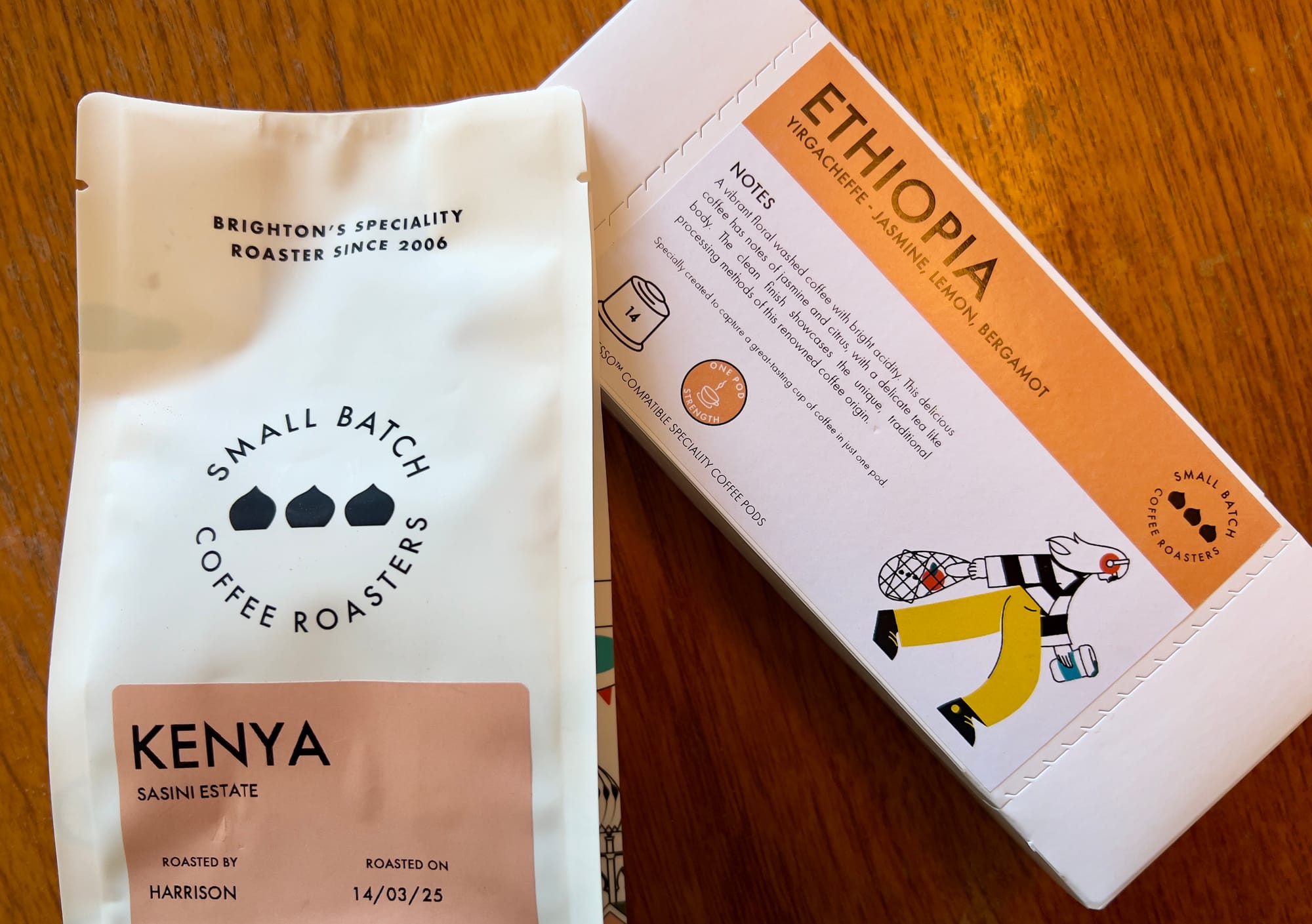 A close-up shot shows two packages of coffee on a wooden surface. On the left is a white bag labeled "KENYA SASINI ESTATE" in a bold, sans-serif pink font. Below this, in smaller black text, it reads "ROASTED BY HARRISON" and "ROASTED ON 14/03/25". Above the Kenya label, the bag features a circular logo with three stylized black coffee beans and the words "SMALL BATCH COFFEE ROASTERS" encircling them.