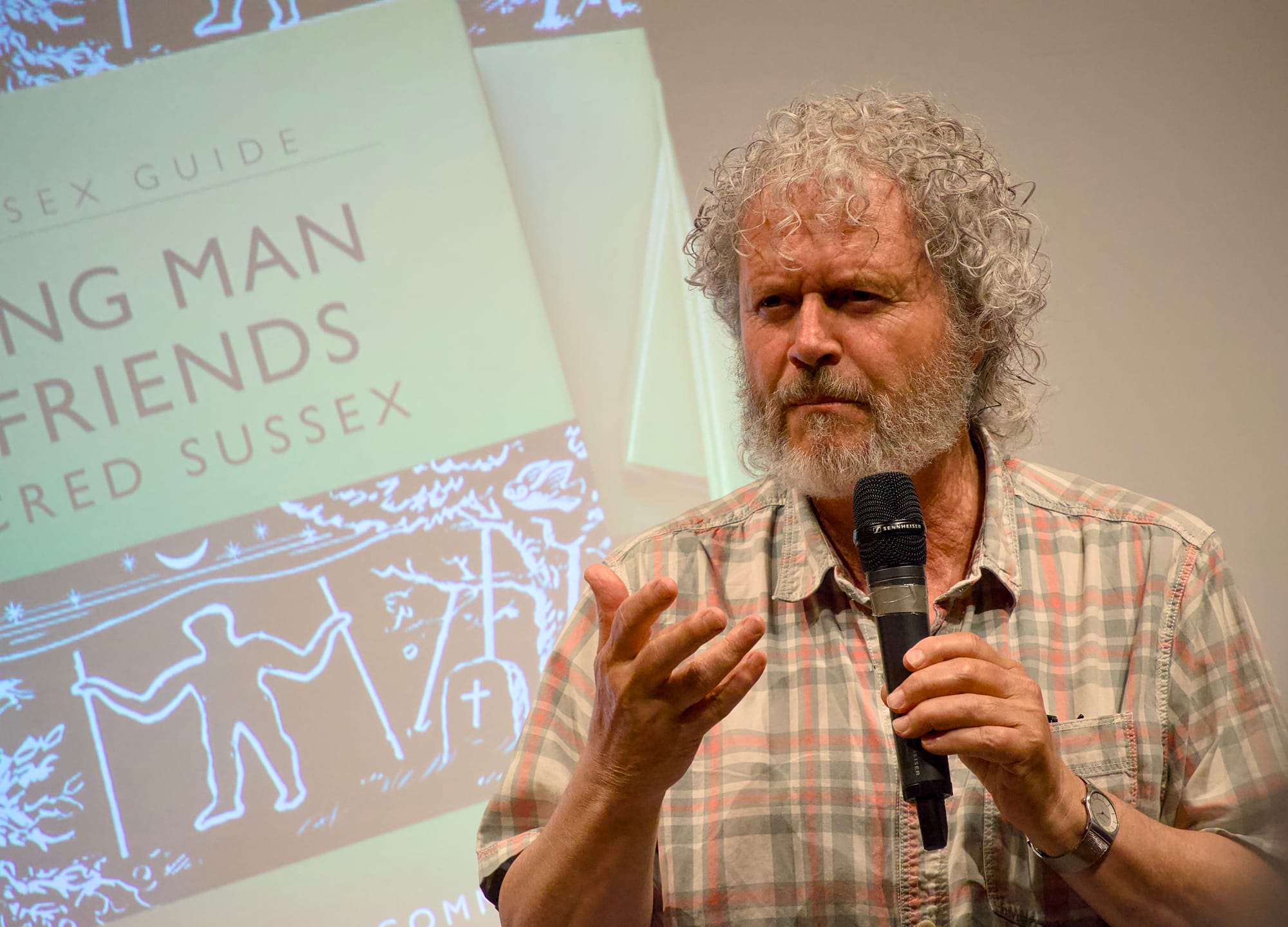 A man with curly gray hair and a beard, wearing a plaid short-sleeved shirt, speaks into a handheld microphone. He gestures with one hand while standing in front of a projection of a book cover titled "LONG MAN & FRIENDS" with the subtitle "SACRED SUSSEX" and illustrated with a figure holding two staffs near a tree and gravestone. 