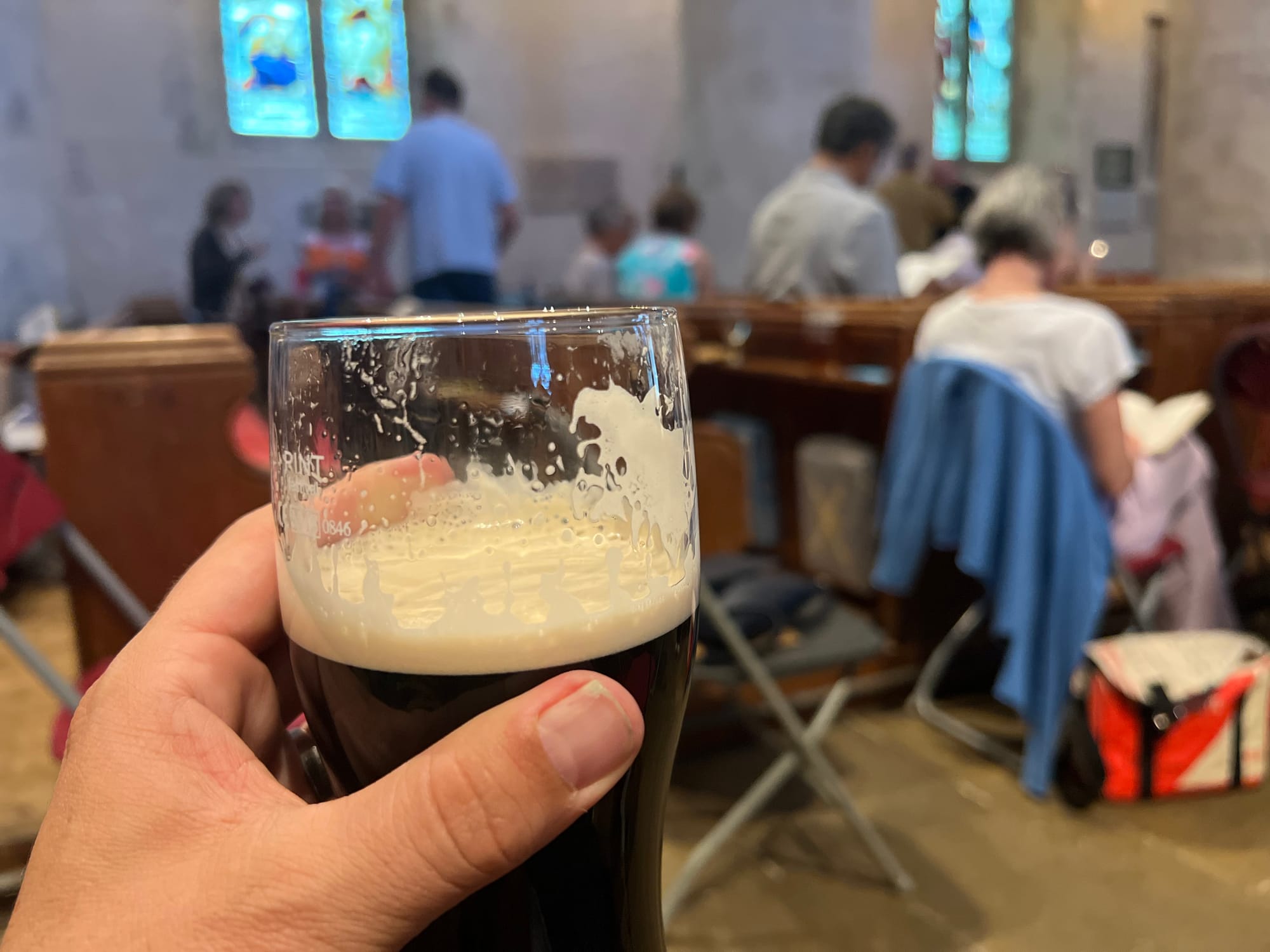 A person holds a pint of dark beer with a creamy head in the foreground, partially consumed, while seated inside a church. The background is softly out of focus, showing people sitting on wooden pews, some reading, with stained glass windows glowing in the stone walls. The scene blends casual socializing with a sacred setting.