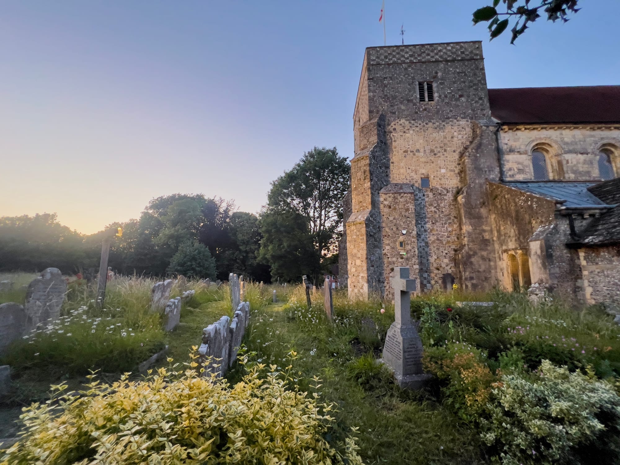 A stone church with a square tower stands in the evening light, surrounded by a peaceful graveyard with weathered headstones and overgrown wildflowers. The sky is clear with a soft gradient from sunset, and tall trees form a natural border behind the churchyard.