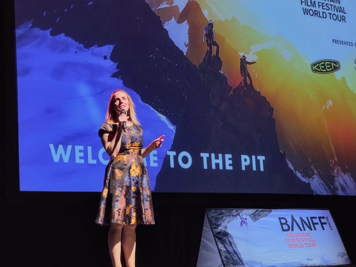 A woman in a floral dress speaks on stage at the Banff Mountain Film Festival, with a screen behind her showing climbers