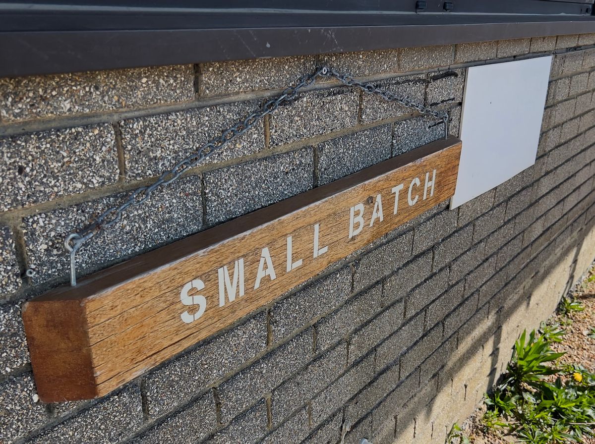 Wooden sign reading “SMALL BATCH” hanging on a brick wall by a metal chain.