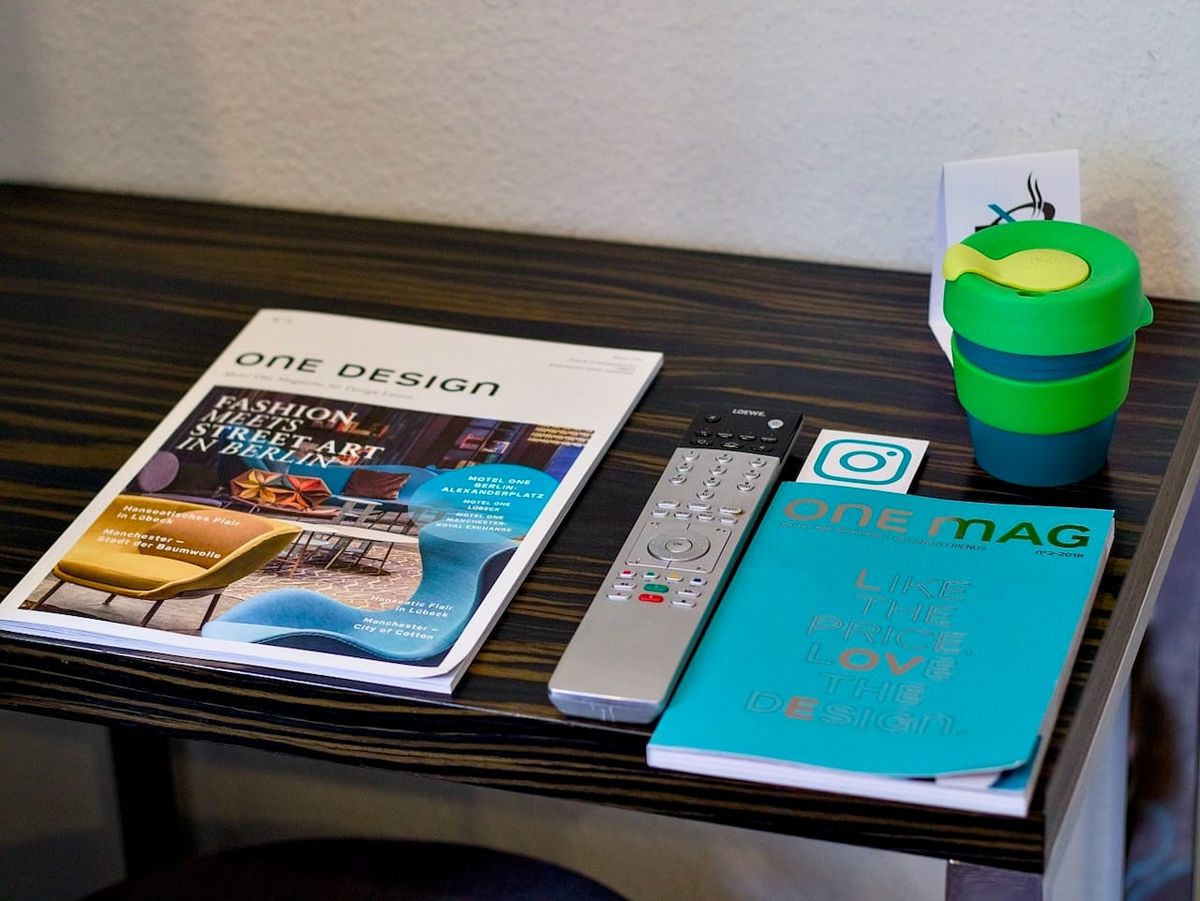 Hotel room side table with design magazines, a TV remote, an Instagram-branded booklet and a reusable coffee cup.