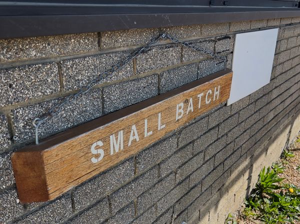 Wooden sign reading “SMALL BATCH” hanging on a brick wall by a metal chain.