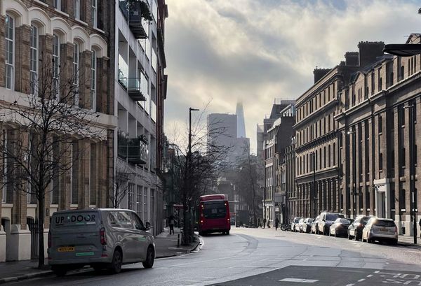 London street scene with brick buildings and parked cars, a red bus driving away, winter light and clouds, and the Shard skyscraper rising in the hazy background.
