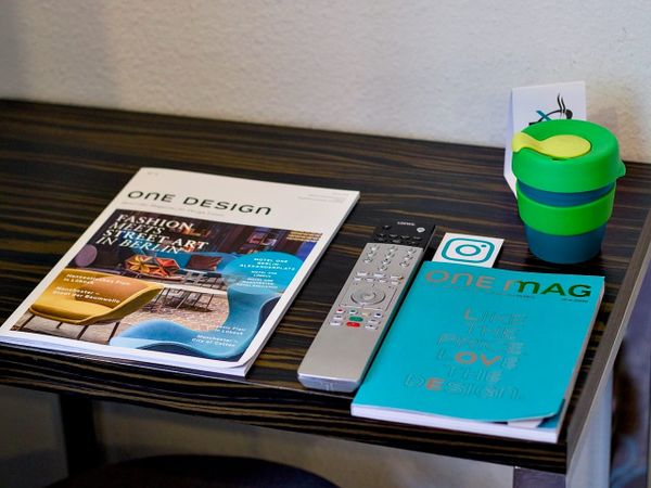 Hotel room side table with design magazines, a TV remote, an Instagram-branded booklet and a reusable coffee cup.