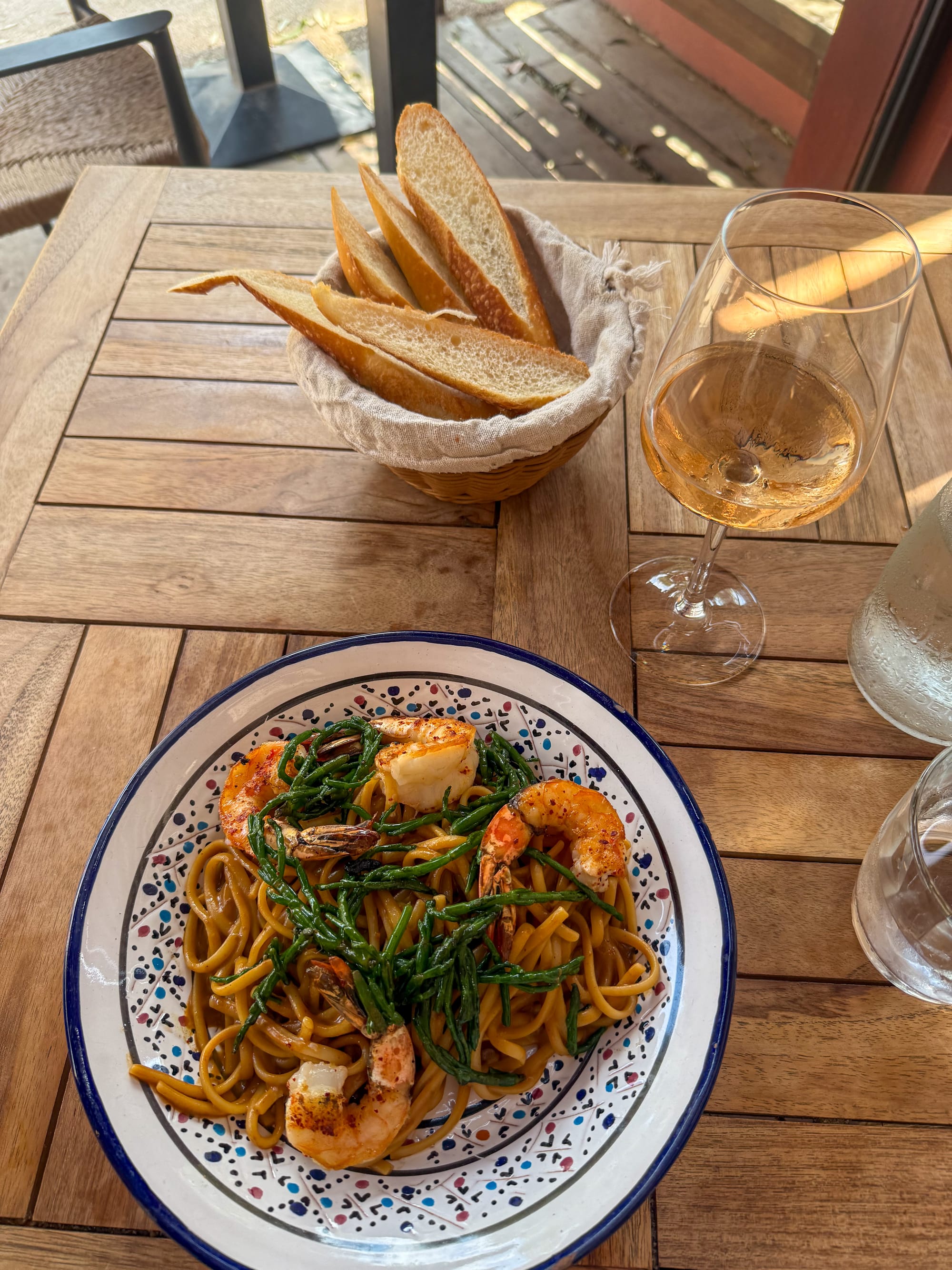 Shrimp pasta with greens, a basket of bread and a glass of rosé on a wooden outdoor table in Porquerolles.
