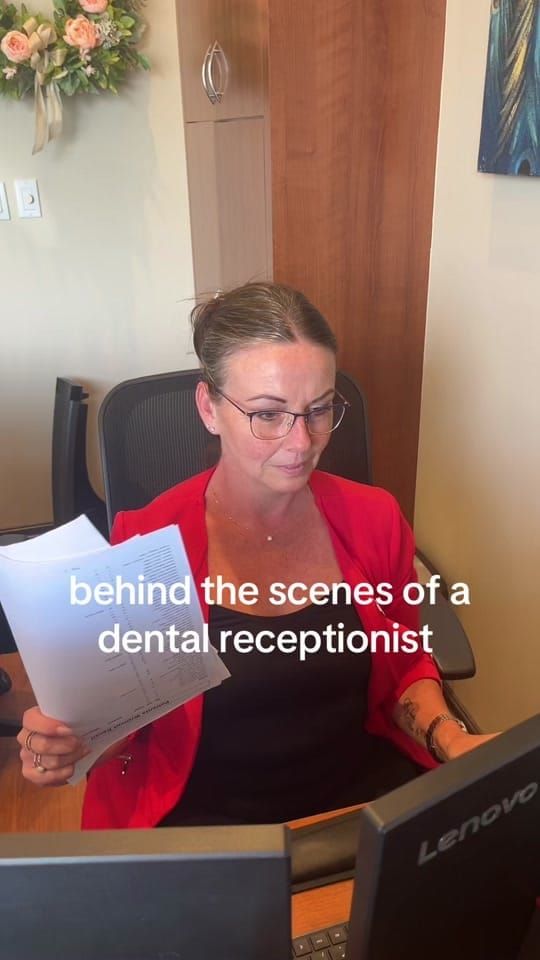 A Longworth Dental Bowmanville receptionist in a red blazer working at the front desk with paperwork, with on-screen text reading: behind the scenes of a dental receptionist.