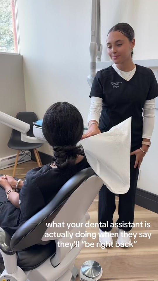 A masked Longworth Dental Courtice assistant stands beside a patient reclined in the dental chair, holding a paper bib.