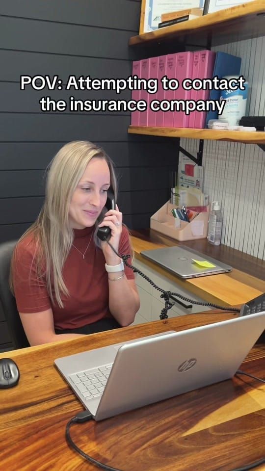 A Longworth Dental Courtice receptionist on the phone at the front desk with a laptop, with on-screen text reading: POV: attempting to contact the insurance company.