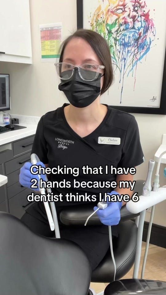 Chelsea, a masked Longworth Dental Courtice assistant, sits in the dental chair holding suction and the air-water syringe.