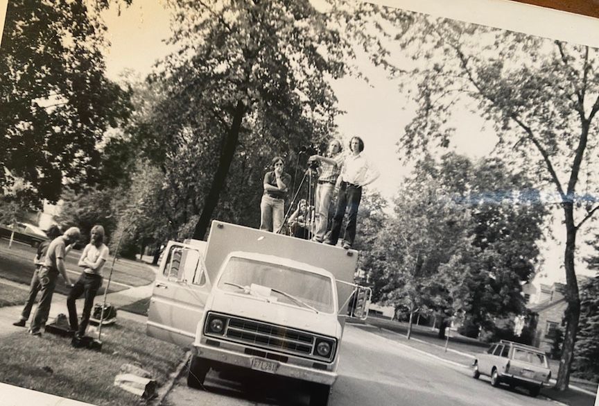 Black & white photo of a film crew on set. Several men standing on top of cargo van w/ a camera.